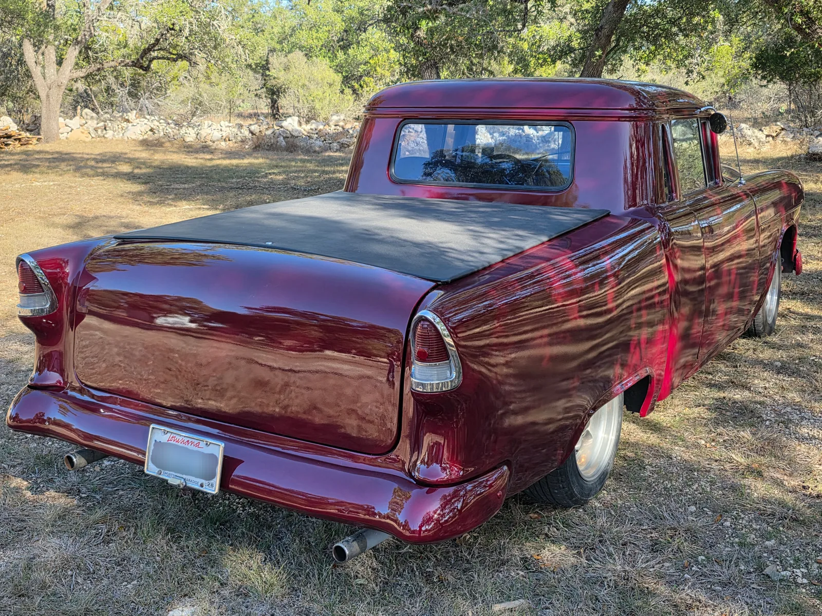 Rear three-quarter view of a classic 1950s pickup conversion with a deep metallic red custom paint finish, parked on grass near trees and a stone wall.