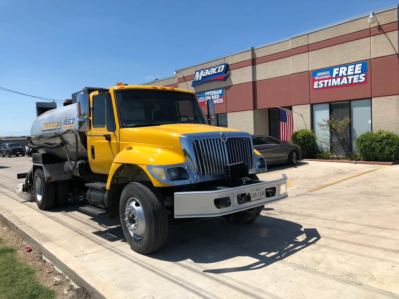 A yellow commercial water tank truck with a chrome tank parked on pavement in front of an auto body shop.